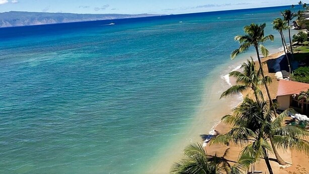 A beach with clear turquoise water, palm trees, and a distant mountain range. - Check out our Summer Discounts and Fantastic Views! (Lahaina)