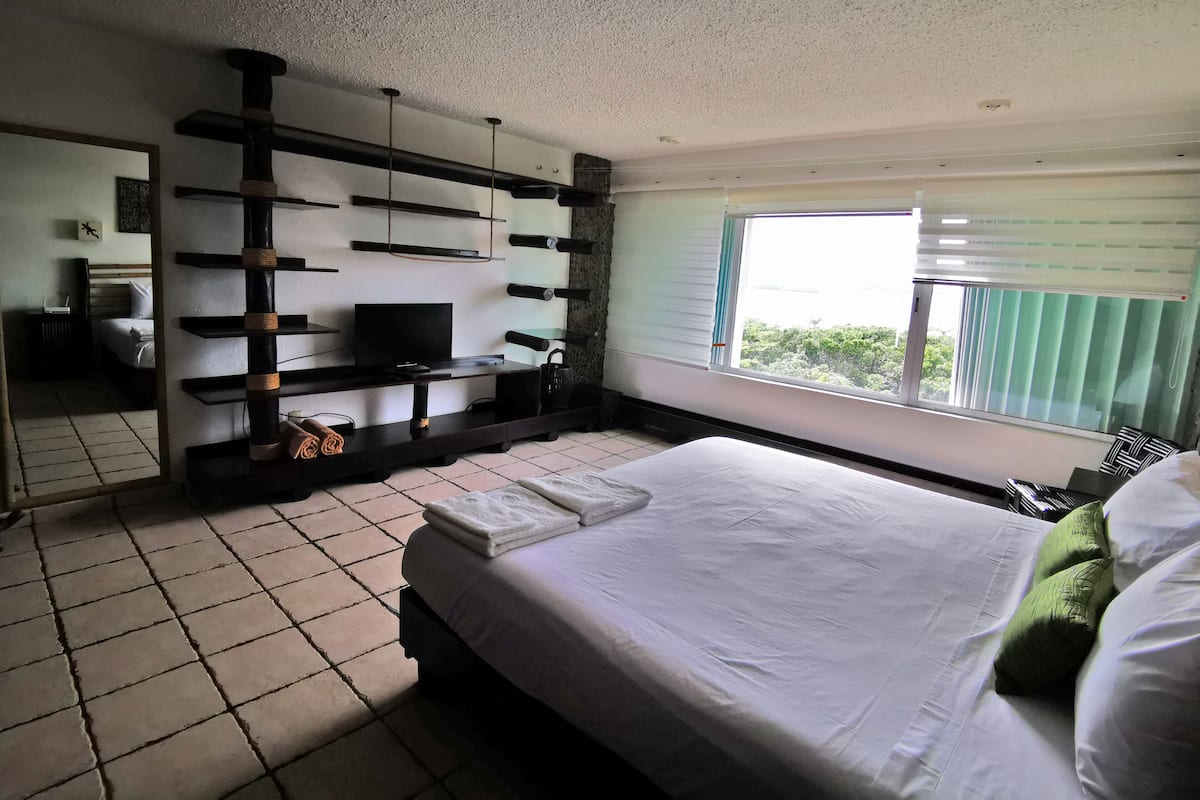 A bedroom with a bed, a TV on a stand, a window with curtains, and a tiled floor.