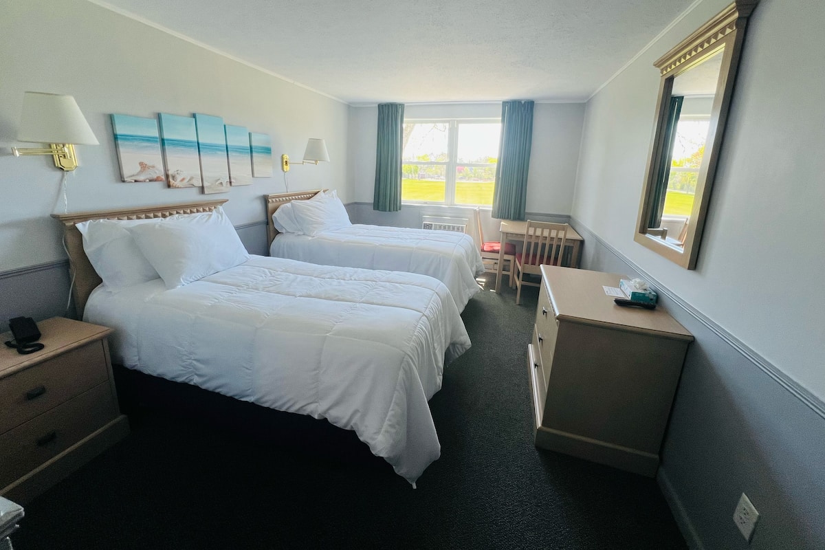 A hotel room with two beds, a desk, a chair, a lamp, and a window with curtains.
