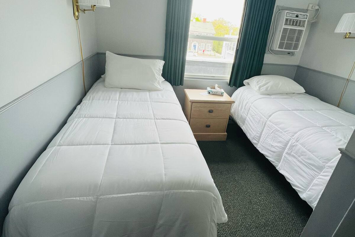 A hotel room with two single beds, a bedside table with a lamp, a window with curtains, and an air conditioning unit.