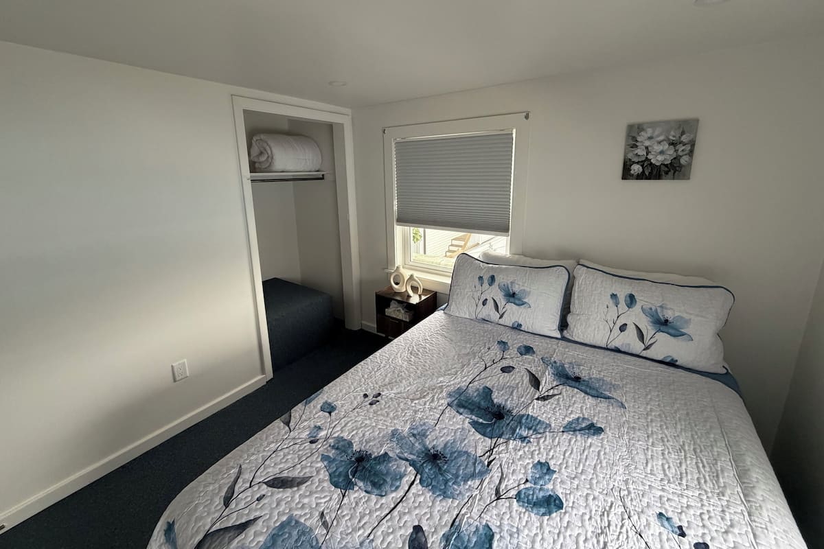 A bedroom with a bed, a window with blinds, a bedside table, a wardrobe, and a small bench.