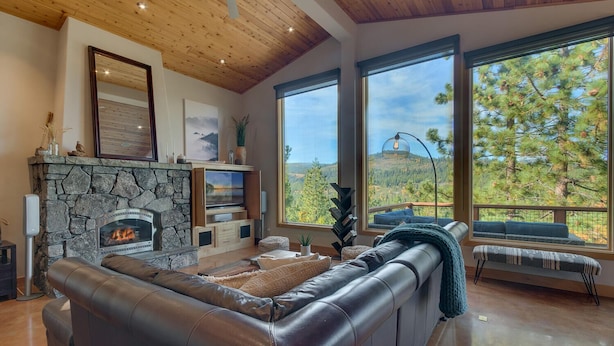 A modern living room with a stone fireplace, leather sofa, and large windows offering a view of trees and hills.