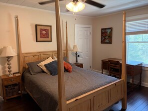 A four-poster bed with a wooden headboard, a bedside table with a lamp, a desk with a chair, and a window with blinds.