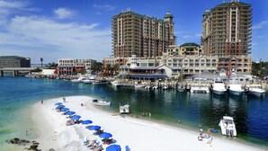 Un hôtel en bord de mer composé de plusieurs immeubles de grande hauteur, un port de plaisance avec des bateaux, et une plage de sable fin équipée de chaises longues et de parasols.