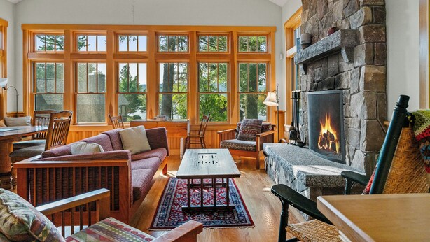 A cozy living room with a stone fireplace, large windows, wooden furniture, and a rug. - Saddleback Lake Lodge 02 | Close to Saddleback (Rangeley)
