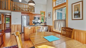 A spacious kitchen with wooden cabinets, a stainless steel refrigerator, and a microwave. There is a dining area with a wooden table and chairs, and a large window providing natural light. - Saddleback Lake Lodge 02 | Close to Saddleback (Rangeley)