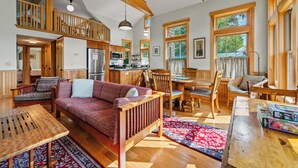 A spacious living area with a high ceiling, wooden beams, a large sofa, a dining table, and a kitchen in the background.