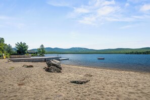 A lakeside beach with a wooden dock extending into the water, surrounded by trees and hills in the background. - Saddleback Lake Lodge 02 | Close to Saddleback (Rangeley)