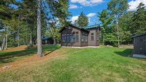 A cabin with a sloped roof, surrounded by a grassy yard and tall trees. - Saddleback Lake Lodge 02 | Close to Saddleback (Rangeley)