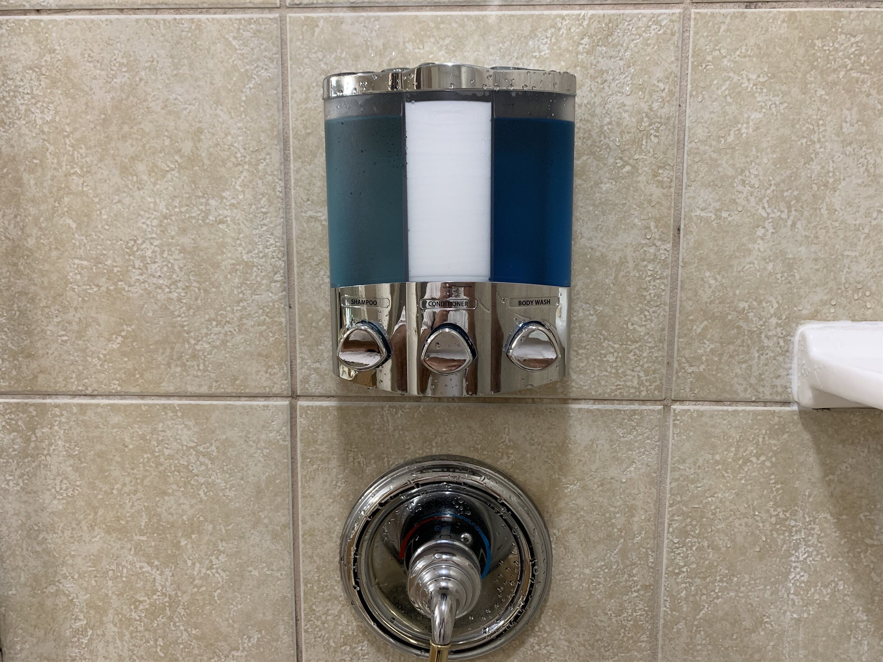 Un distributeur mural doté de trois robinets étiquetés « shampoing », « après-shampoing » et « gel douche ».
