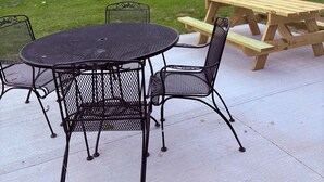 Um pátio externo com uma mesa redonda de metal e cadeiras, uma mesa de piquenique de madeira e uma área gramada com árvores ao fundo.