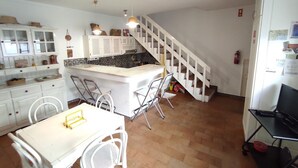 Une cuisine avec des meubles blancs, une table à manger avec des chaises, un petit îlot central et un escalier.