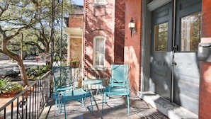 A covered porch with two chairs and a small table, a brick building, and a dark-colored door.