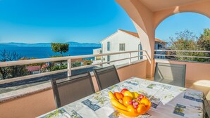 Un balcone con un tavolo apparecchiato per un pasto, che si affaccia su una vista costiera con una casa e montagne sullo sfondo.