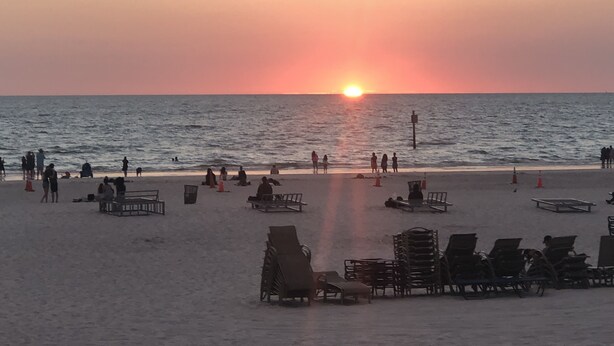 Una playa con gente tomando el sol y sillas de playa, un atardecer al fondo y un mar tranquilo.