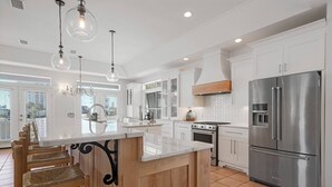 A modern kitchen with white cabinetry, a stainless steel refrigerator, a wooden island with bar stools, and pendant lighting. - Endless Summer - Home in Prestigious Community with Gulf Views & Secluded Pool Area (Miramar Beach)