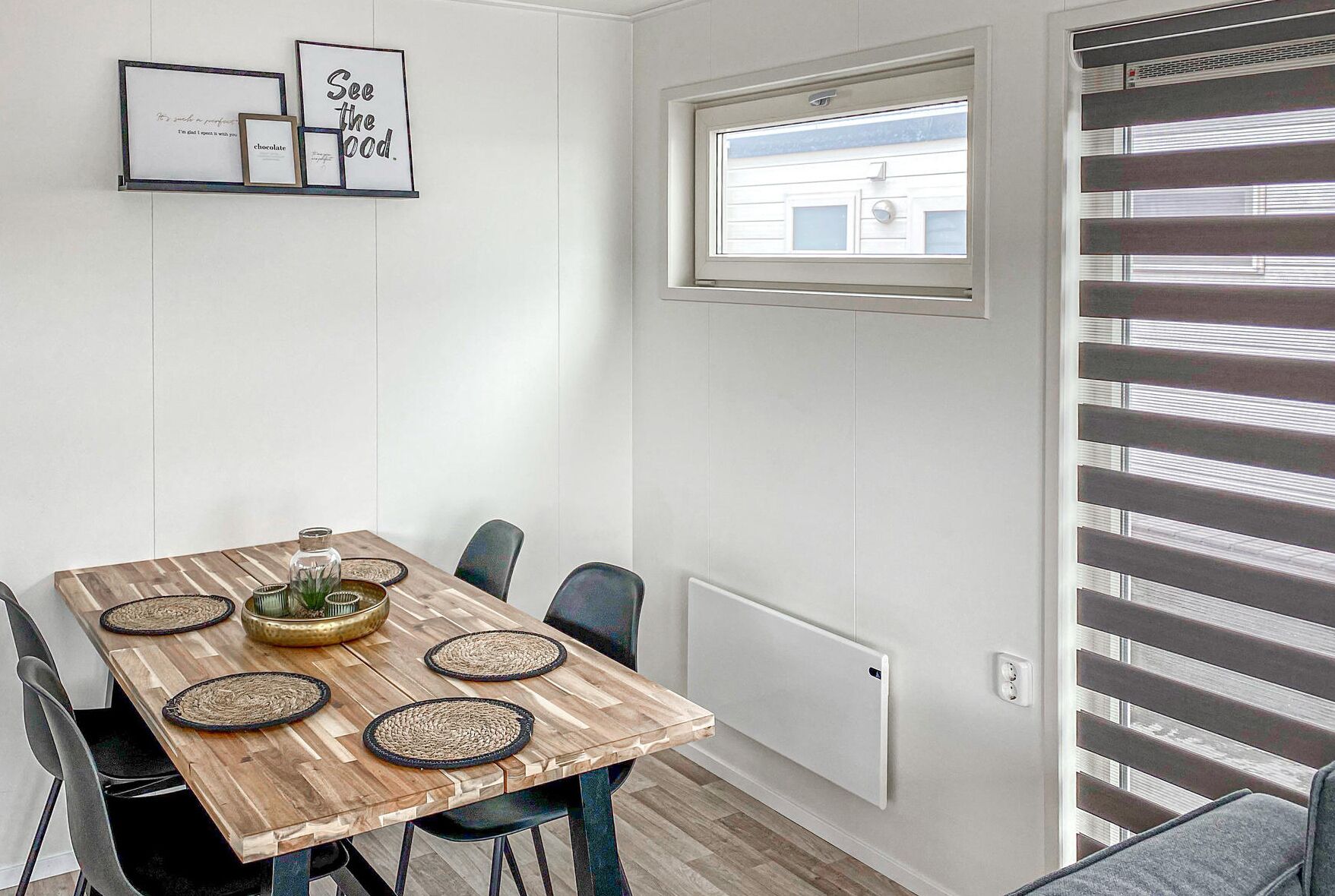 Un comedor con una mesa de madera, individuales tejidos y un pequeño centro de mesa con planta. Hay dos letreros enmarcados en la pared y una ventana con persianas.