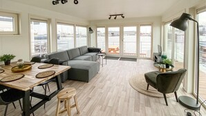 En moderne stue med en grå hjørnesofa, et træspisebord med vævede underlag, et rundt sofabord og en sort lænestol. Store vinduer giver udsigt til vandet og kajen udenfor.