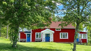 Ett rött hus med en vit veranda och blå dörrar, omgivet av grönt gräs och träd.