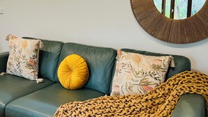 A green leather sofa with floral and yellow cushions, a woven throw blanket, and a round mirror on the wall. - The Yellow Feather Cottage - Enchanting & Private Romantic Stay! (Wentworth Falls)