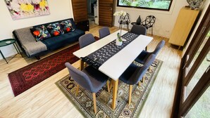 A modern living room with a dining area, featuring a sofa, a dining table, and a large window with a view. - The Yellow Feather Cottage - Enchanting & Private Romantic Stay! (Wentworth Falls)