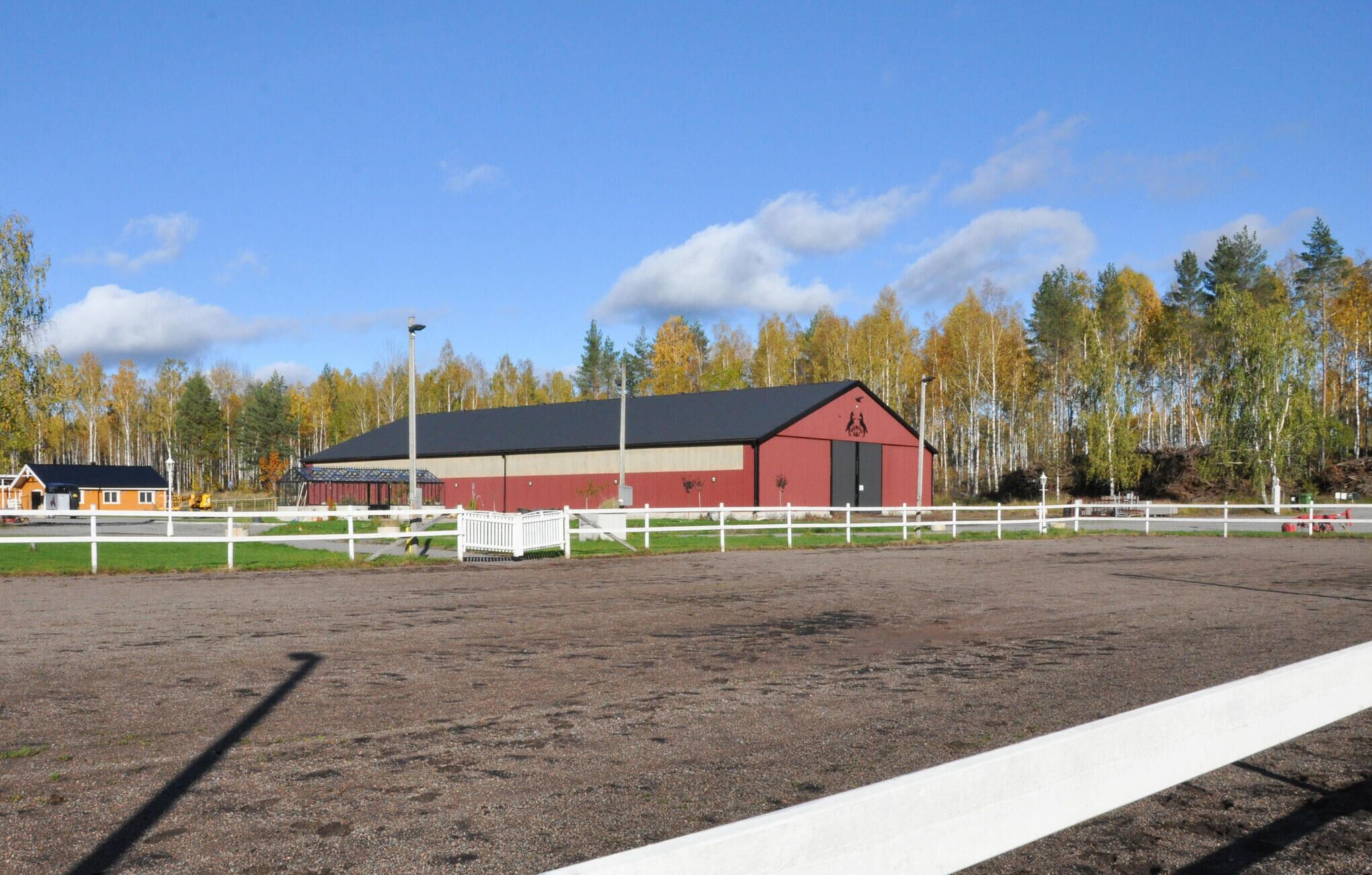 A stable with a red and white exterior, surrounded by a white fence, set against a backdrop of trees and a clear sky.