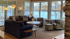 Ein gemütliches Wohnzimmer mit einem dunkelblauen Sofa, gemusterten Sessel und einem runden Couchtisch. Große Fenster bieten Blick auf einen See und Bäume.