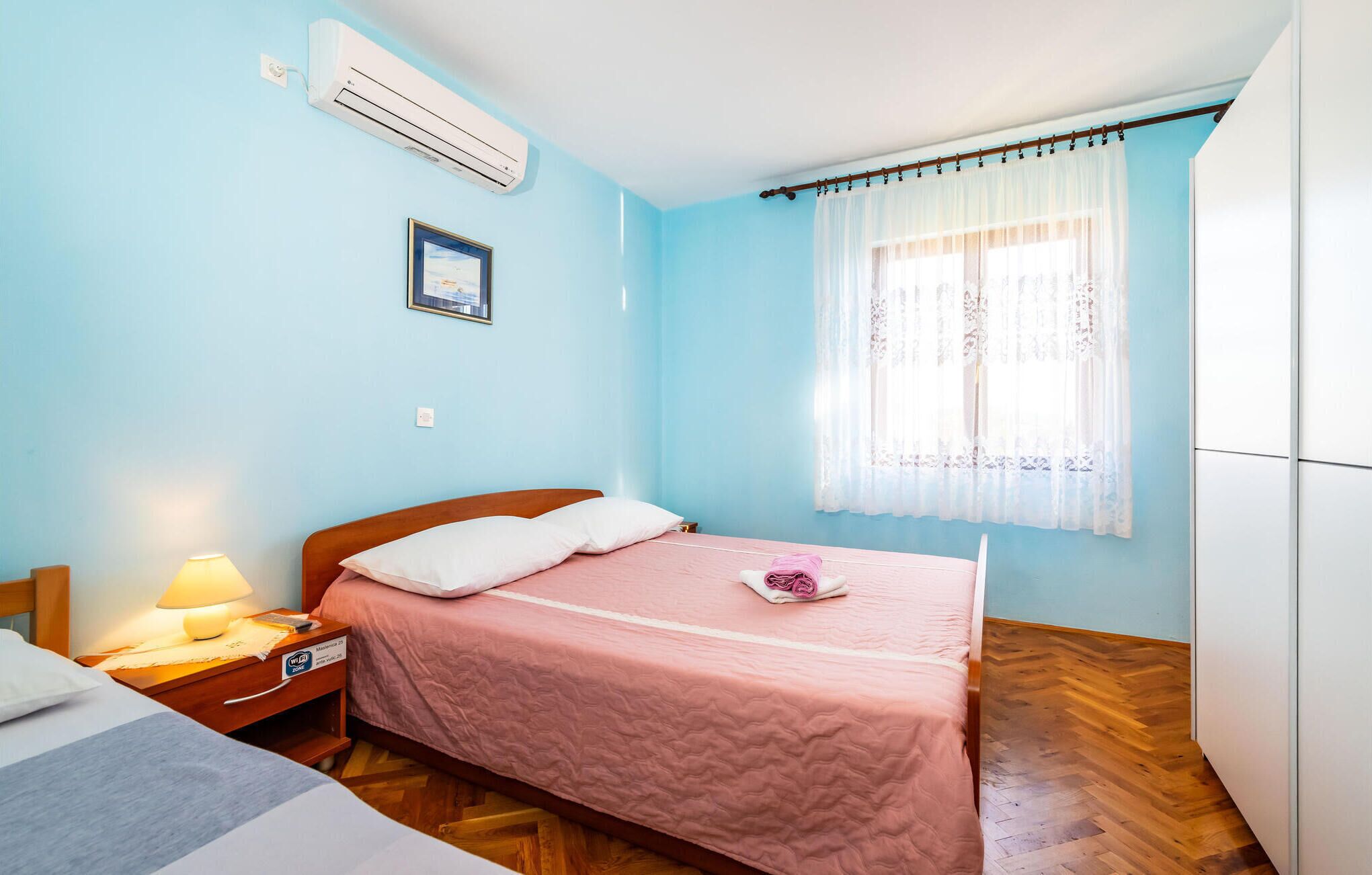 A bedroom with a bed, a nightstand, a lamp, a window with sheer curtains, and an air conditioning unit.