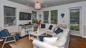 A modern living room with a white sofa, blue armchair, and a coffee table. There is a TV mounted on the wall, and the room has large windows with blinds. - Luxury Getaway on Bald Head Island - 5-Star Property - Discounts for September (Bald Head Island)