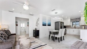 Ein modernes Wohnzimmer mit Sofa, Hocker, Esstisch und Stühlen. Es gibt eine Küchenzeile mit weißen Schränken und einem Kühlschrank.