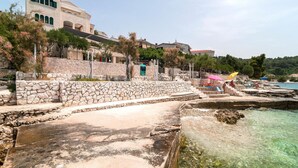Zona costera con escalones de piedra que bajan a una playa, agua cristalina y edificios con balcones.