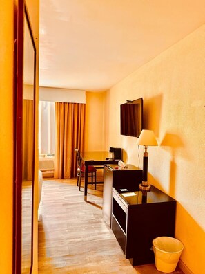 A hotel room with a desk, chair, TV, lamp, and a trash bin.