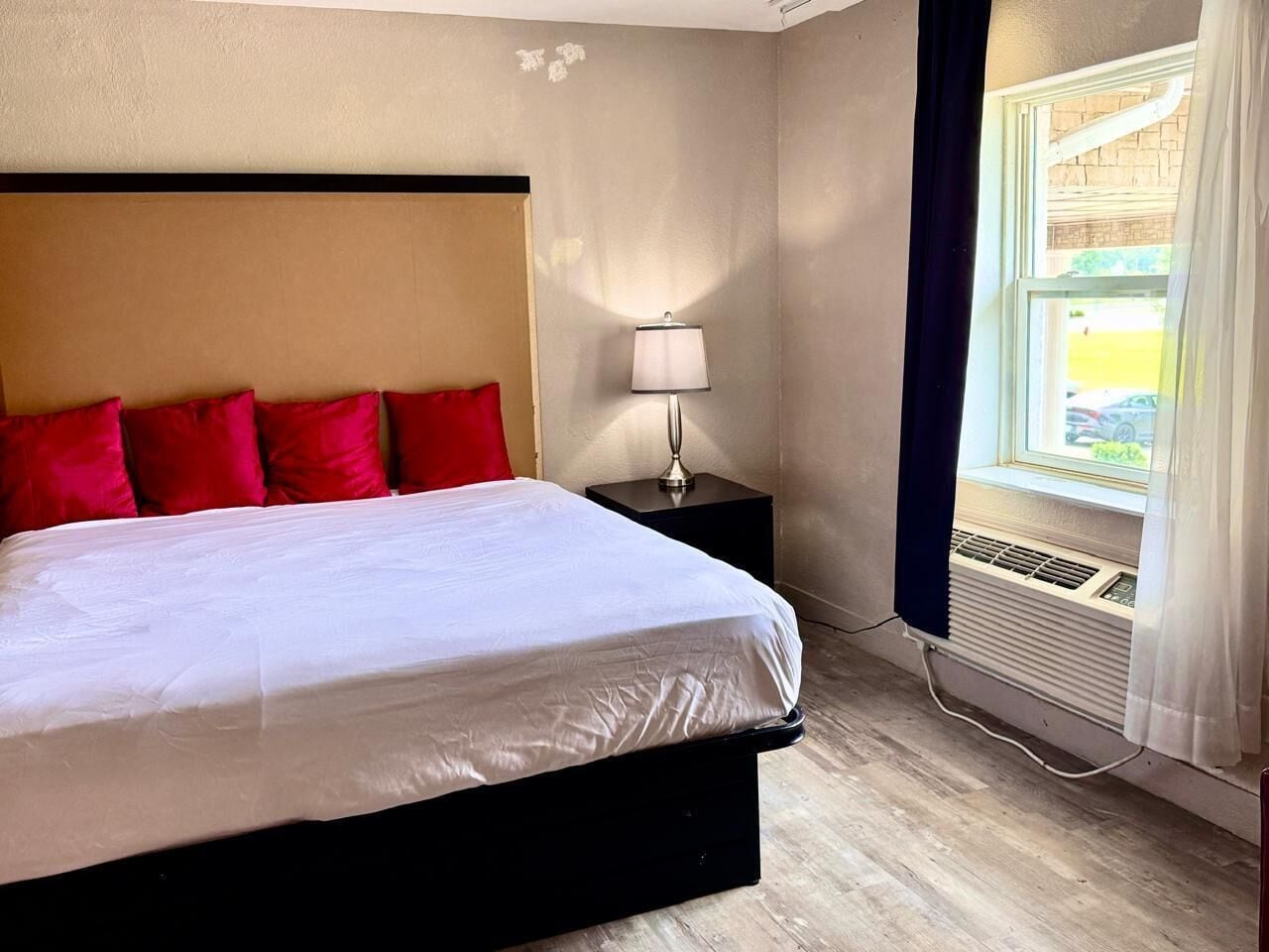 A hotel room with a bed, red pillows, a nightstand with a lamp, and a window with curtains.