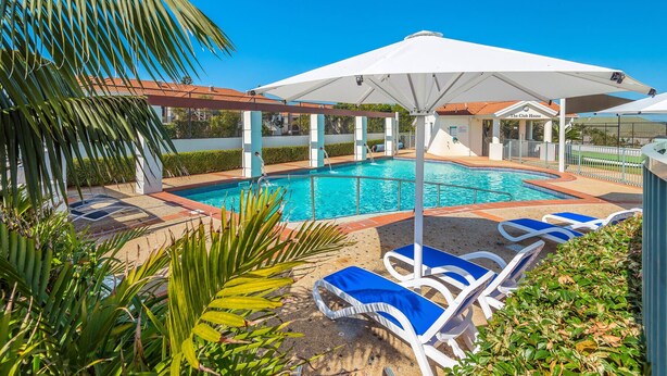 Une piscine entourée de palmiers, agrémentée de chaises longues et de parasols, et à proximité d’un bâtiment portant l’inscription « The Club House ».