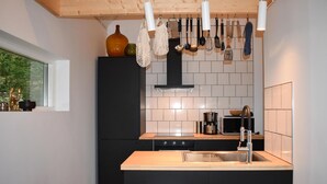 Une cuisine de taille réduite avec un réfrigérateur noir, un four à micro-ondes et un évier. Des ustensiles de cuisine sont suspendus au-dessus de la cuisinière, et une fenêtre se trouve sur le côté gauche.
