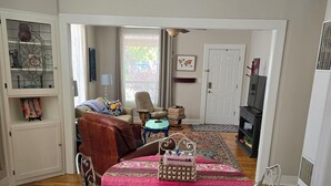 Ein gemütliches Wohnzimmer mit Holzboden, einem Esstisch mit floralem Tischläufer, einem braunen Ledersofa, einem beigefarbenen Sessel und einem Kamin.
