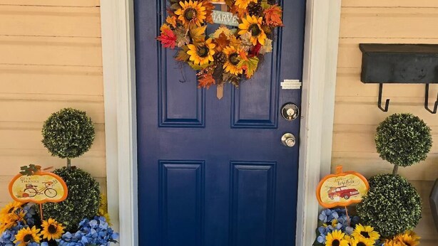 Eine blaue Tür mit einem Kranz, zwei Kübelpflanzen mit Sonnenblumen und Hortensien sowie je ein Schild auf beiden Seiten.