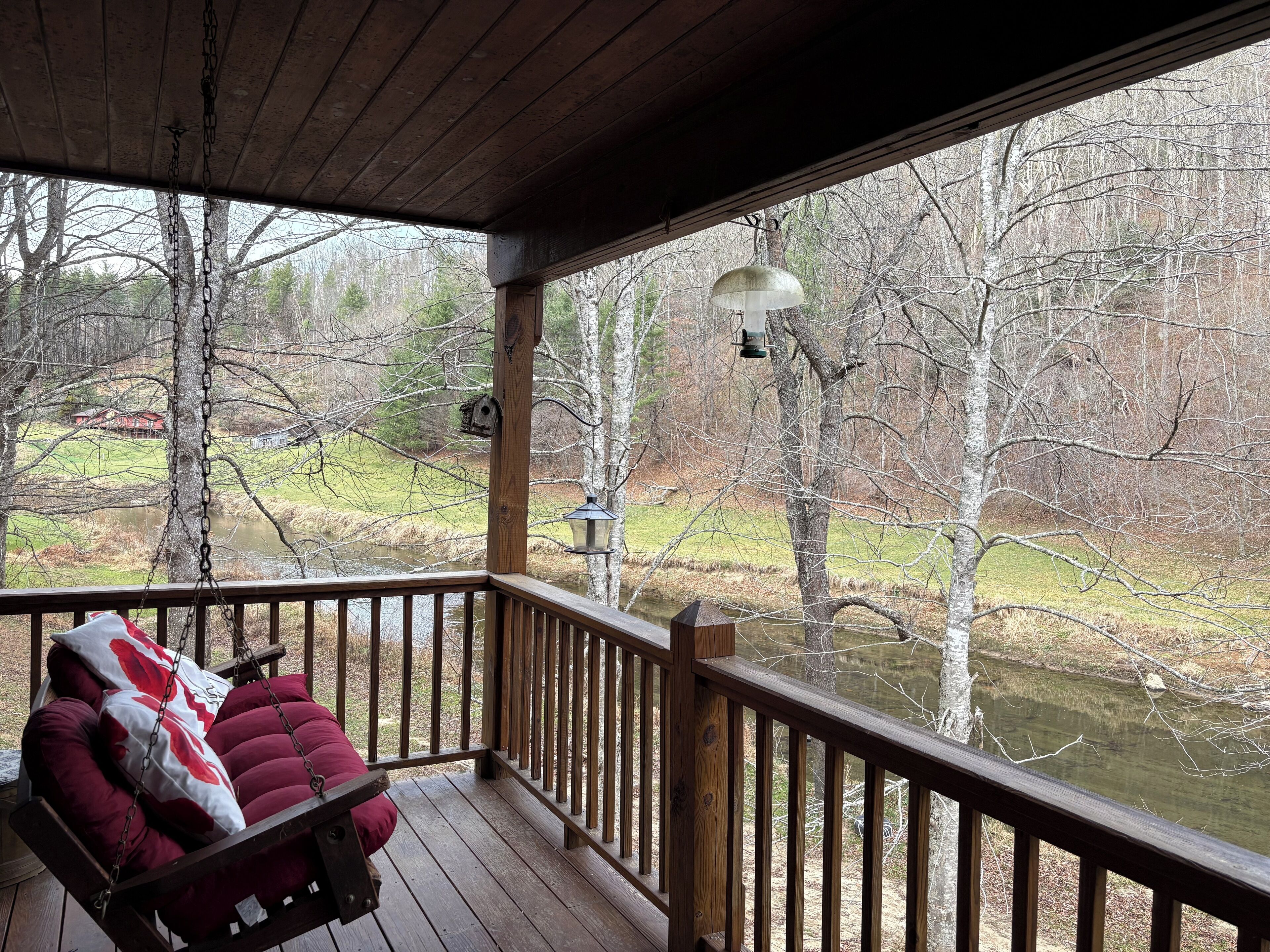 A wooden deck with a swing chair and a view of a lake surrounded by trees.