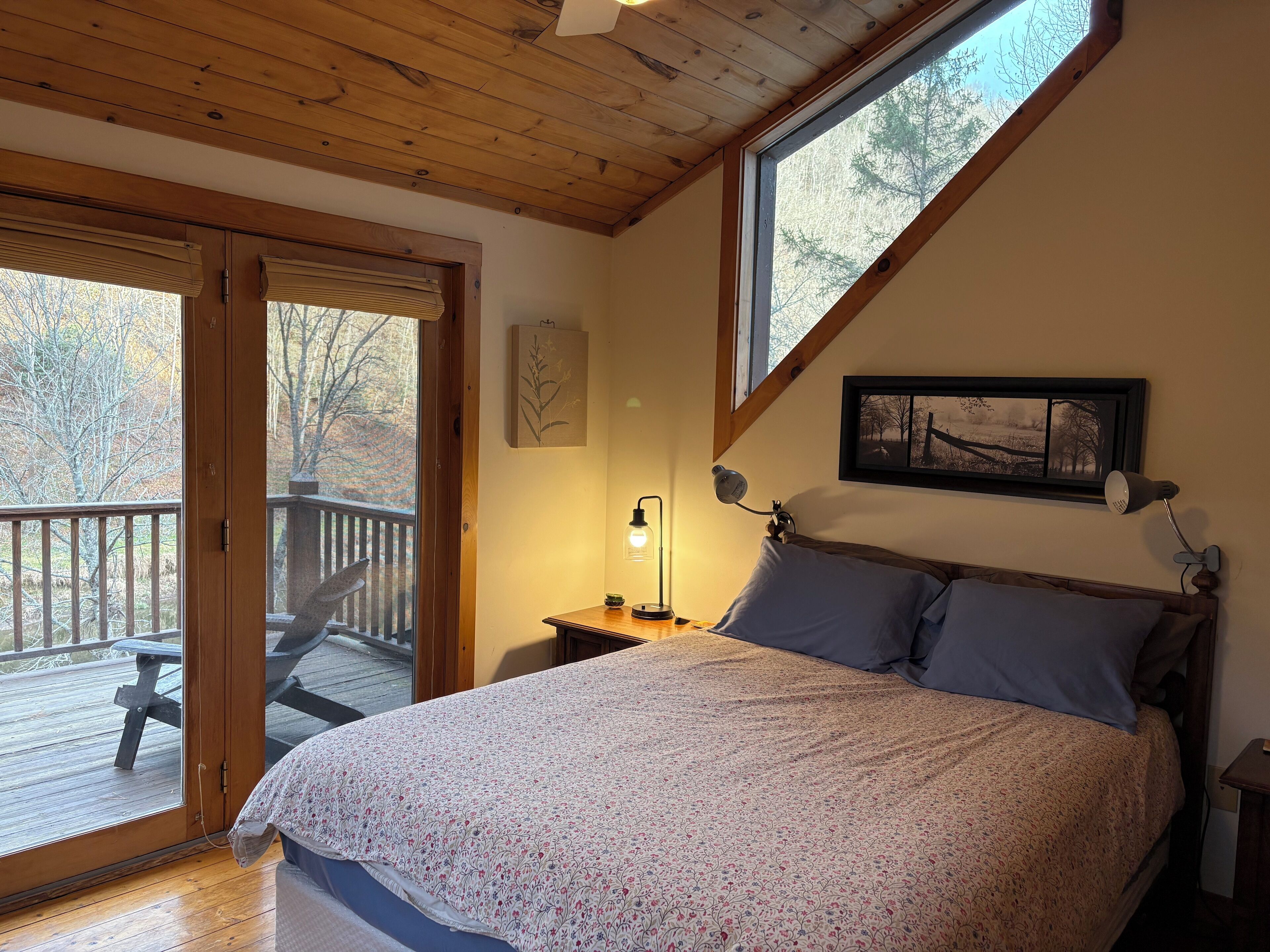 A bedroom with a wooden ceiling, a bed with a floral bedspread, a nightstand with a lamp, and a window with a view of trees.