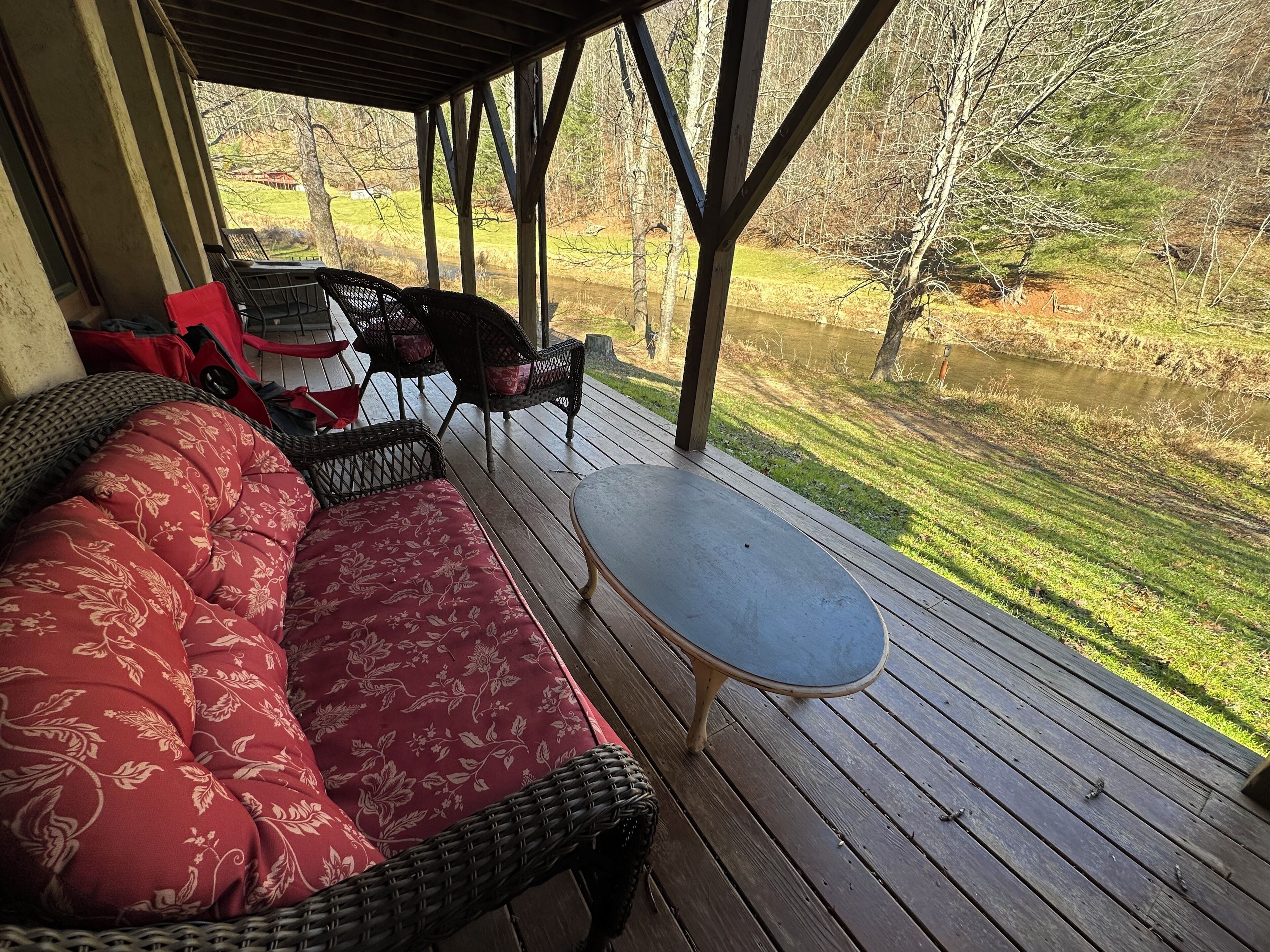 A covered outdoor seating area with a sofa and chairs, a small table, and a view of a pond and trees.