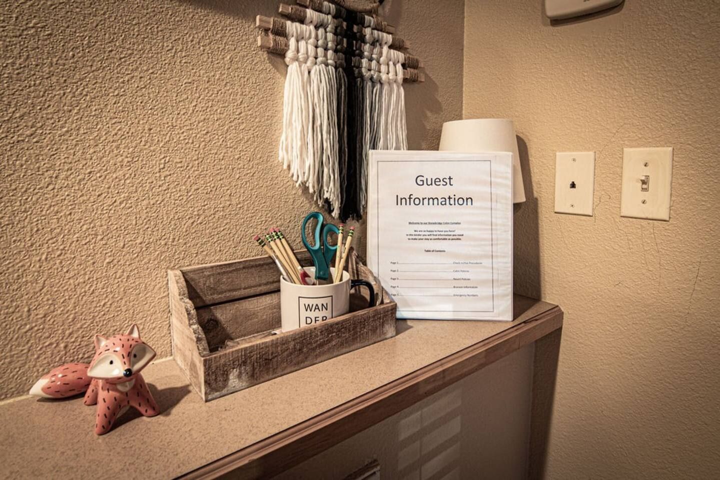 A reception desk with a 'Guest Information' sign, a decorative wall hanging, a wooden box with writing utensils, and a small fox figurine.