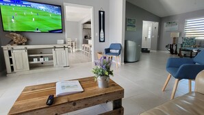 Un salon avec un téléviseur diffusant un match de sport, une table basse en bois sur laquelle se trouvent une télécommande et un livre, des fauteuils bleus et un canapé.