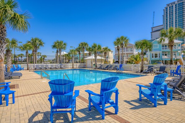 A pool area with blue Adirondack chairs, palm trees, and a clear blue sky.
