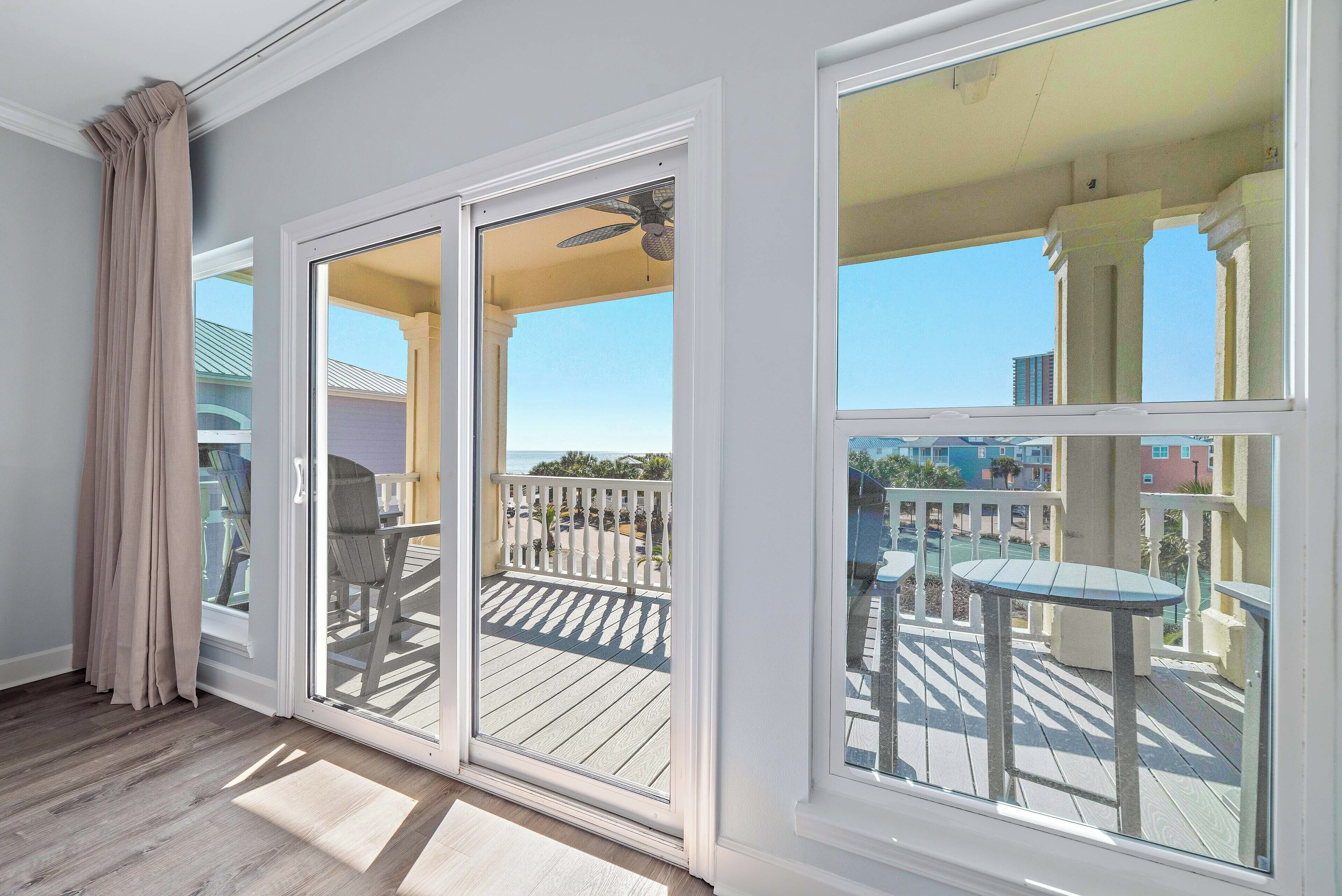 Una habitación con puerta corrediza de vidrio que da a una terraza exterior con ventilador de techo, vista de un edificio más allá de la terraza, y una pequeña mesa y sillas en la terraza.