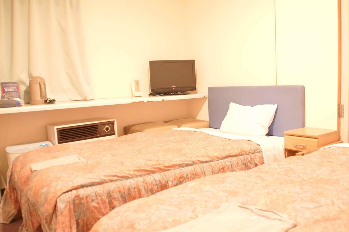 A hotel room with two beds, a television on a shelf, a bedside table, and a window with curtains.