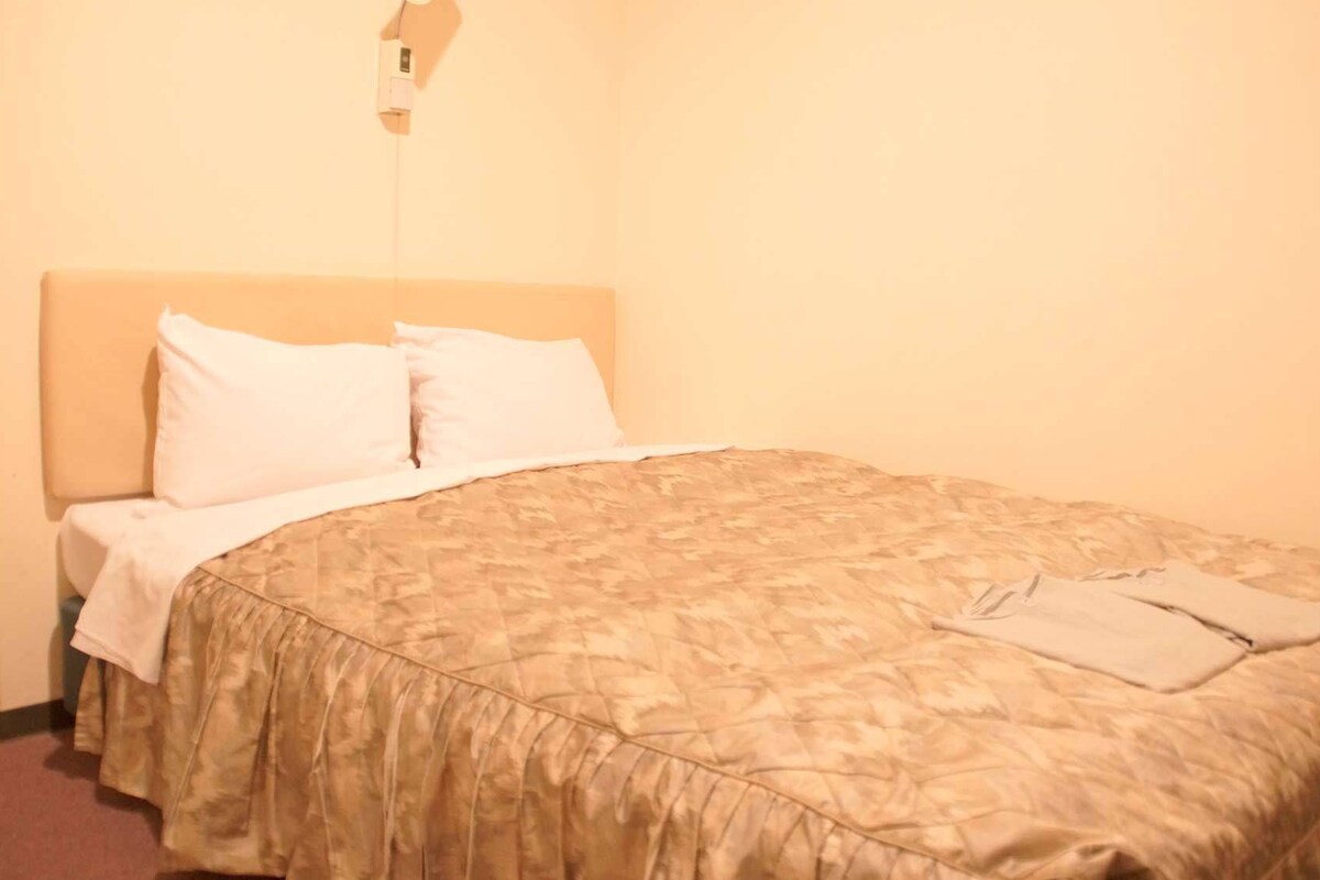 A neatly made bed with a patterned bedspread and white pillows in a room with a plain wall and a light switch above the bed.