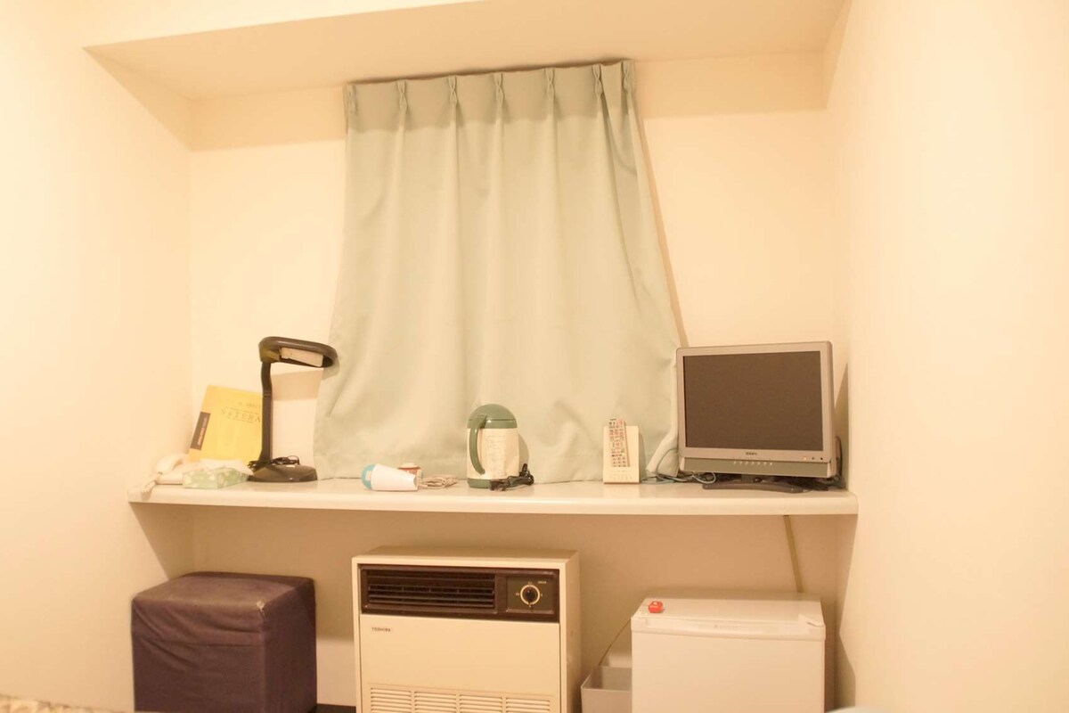 A small, well-lit room with a desk, a computer, a heater, and a window with a curtain.