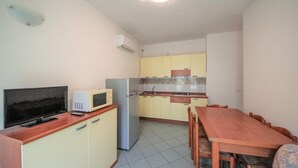 A compact kitchen with a refrigerator, microwave, and stove. There is a dining table with chairs and a television on a cabinet. - Stunning apartment in Rosolina Mare (Rosolina Mare)