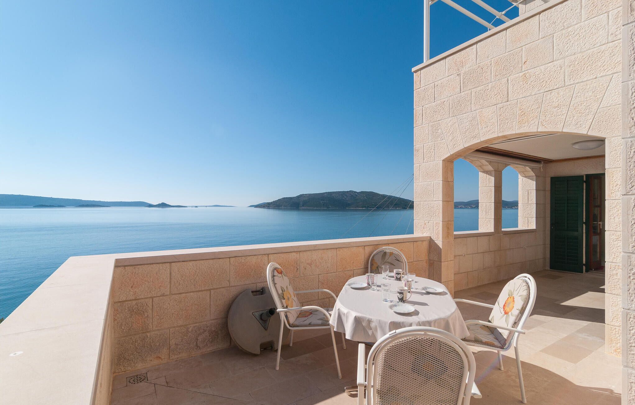 Terraço com mesa posta para dois, com vista para um mar tranquilo e colinas ao longe.