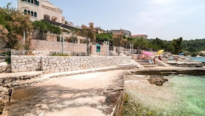 Zona costera con escalones de piedra que bajan a una playa, agua cristalina y edificios con balcones.